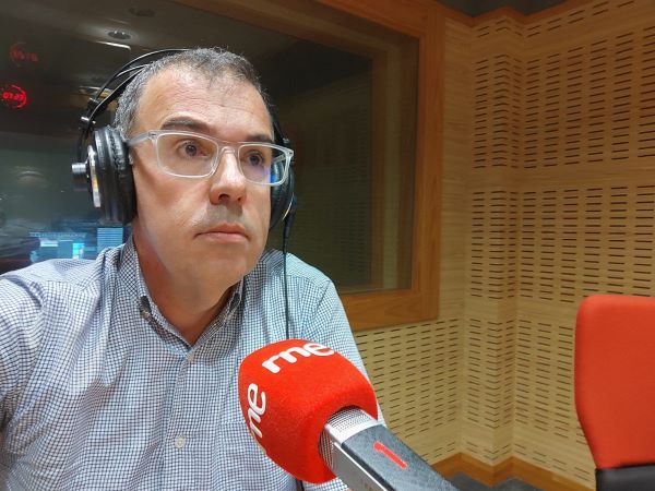 Un hombre de mediana edad con gafas y camisa azul cielo en un estudio de radio. Un micrófono rojo lleva escrito RNE