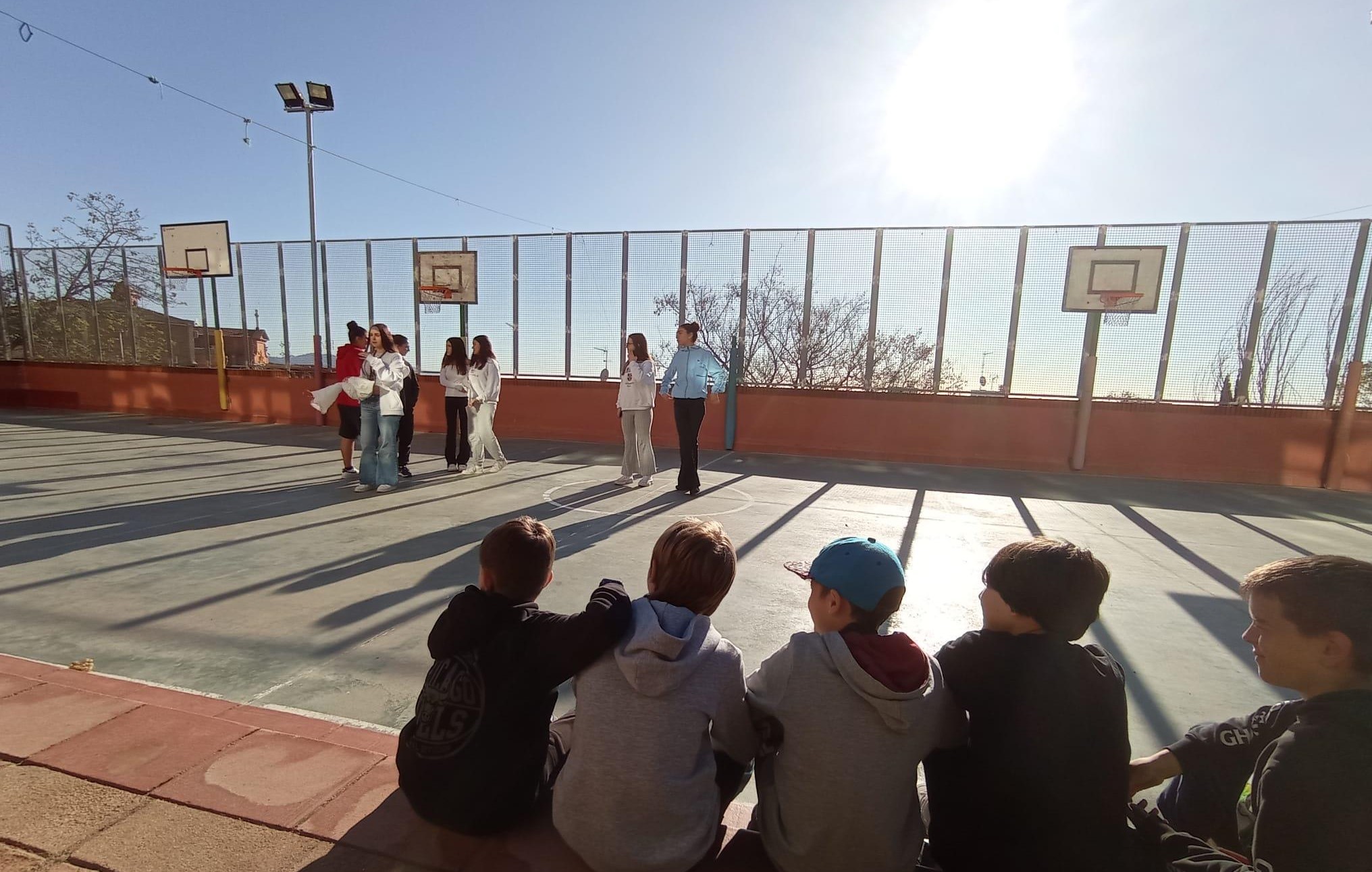 Un grupo de niños observa sentado a un grupo de adolescentes en espacio deportivo al aire libre