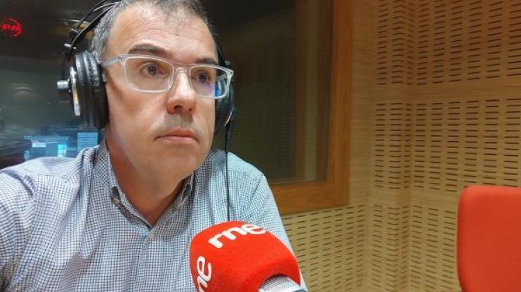 Un hombre de mediana edad con gafas y camisa azul cielo en un estudio de radio. Un micrófono rojo lleva escrito RNE