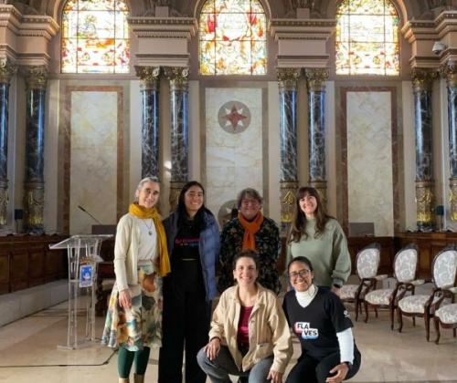 Un grupo de 6 mujeres posa mirando a cámara en un salón señorial. Se ven sillas blancas a la derecha y tres vitrales artísticos en las ventanas del fondo