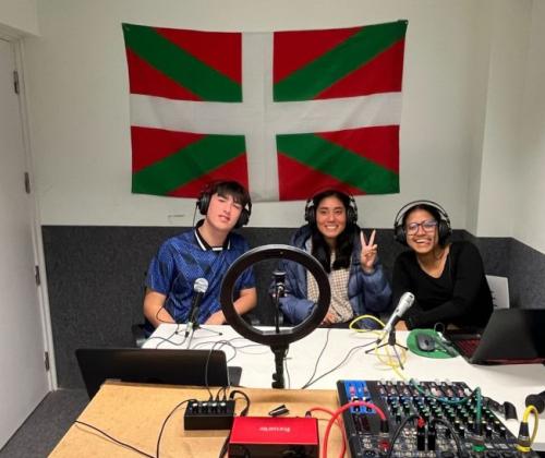 Un grupo de dos chicas y un chico posan sentados en un estudio de radio. Llevan auriculares y al fono se ve la ikurriña, la bandera del país vasco,  con sus colores verde, blanco y rojo